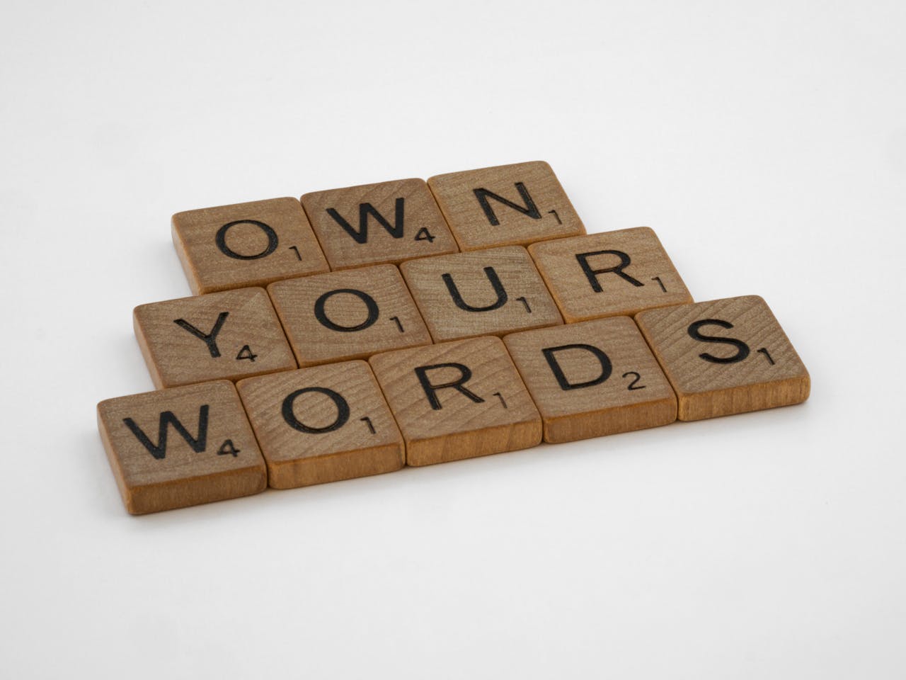 Wooden Scrabble tiles spelling 'Own Your Words' on a white background, symbolizing communication integrity.